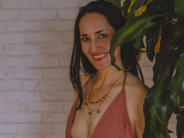 MiaMott with dark hair, red lipstick, smiling, wearing a low-cut, pink top, and gold necklace, standing next to a green plant, showing her tits.