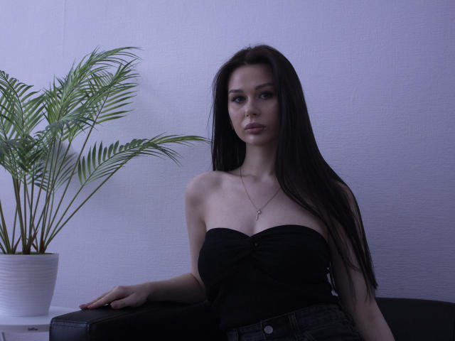 LaylaMolligan with long dark hair, wearing a strapless black top, showing her tits, sitting with a hand on a black couch, potted plant to her left.
