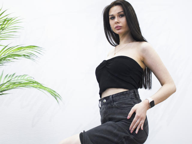 LaylaMolligan has long black hair, a strapless black top, and dark denim shorts, with a watch on her left wrist and a green plant on the left.