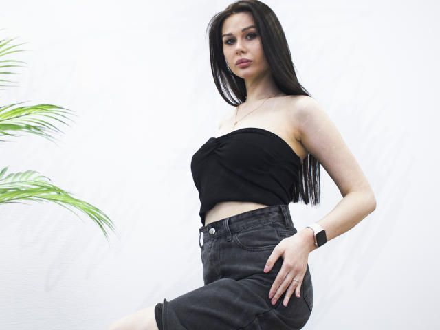 LaylaMolligan with long black hair, strapless black top, and high-waisted black denim shorts, wearing a smartwatch, standing with a hand on her thigh, palm leaves in background.
