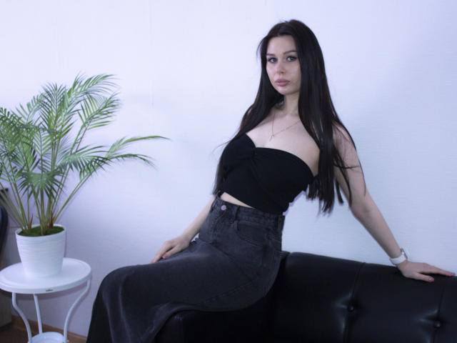 LaylaMolligan with long black hair, wearing a strapless black top and high-waisted dark jeans, sitting on a black leather couch, next to a potted plant on a white table.