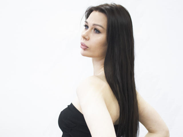 LaylaMolligan with long black hair, pale skin, and blue eyes, wearing a black strapless top, looking over her shoulder with a slight pout. Boobs visible, hands on hips.