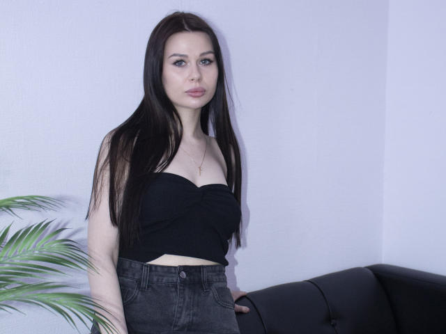 LaylaMolligan with long black hair, wearing a strapless black top and high-waisted black jeans, standing next to a black couch with a green plant in the background.