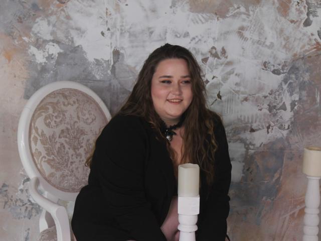 DaniellaGraceZZ with long brown hair, smiling, wearing a black top, holding a white candle, sitting in front of a white chair with pink floral upholstery, on a textured wall.