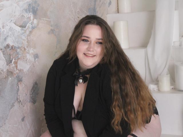 DaniellaGraceZZ with long brown hair, black shirt, and black choker, smiling, showing her tits, sitting, surrounded by white candles.