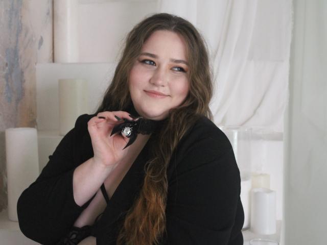 DaniellaGraceZZ with long brown hair, smiling, wearing a black top, holding a black choker with a silver ring, showing off her tits, in a softly lit room.