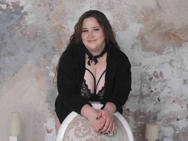 DaniellaGraceZZ with long brown hair, black lace bra, and blazer, leaning on a white chair, smiling, hands on top, wearing black choker with pendant, pale skin, green nails.