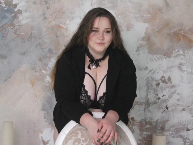 DaniellaGraceZZ with long brown hair, black lace bra, and blazer, showing her big tits, leaning on a white chair, black choker with bell, and painted nails.