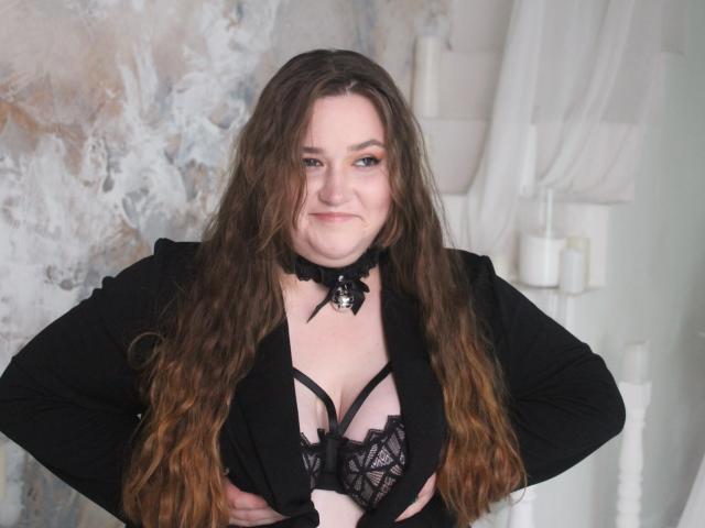 DaniellaGraceZZ with long brown hair, black choker, black jacket, and black lace bra showing her tits, hands on hips, slight smile, white candles in background.