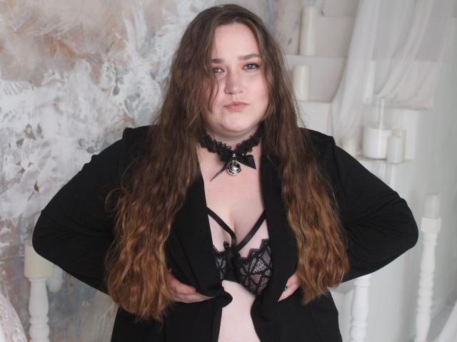 DaniellaGraceZZ with long brown hair, black choker, and black jacket, exposing her black lace bra and pink skin, standing with hands on her jacket, pouty expression.