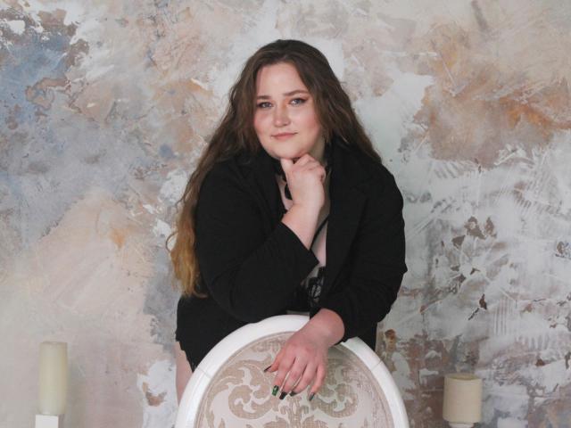 DaniellaGraceZZ with long brown hair, black jacket, and black bra, leaning on a white chair with floral pattern, smiling, hand on chin, candles on either side.