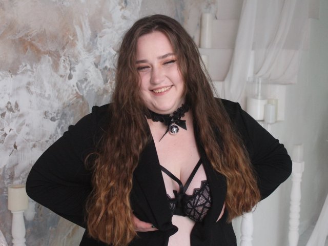 DaniellaGraceZZ with long brown wavy hair, smiling, wearing a black choker, open black jacket, and black lace bra, showing her tits, hands on her hips, in a room.