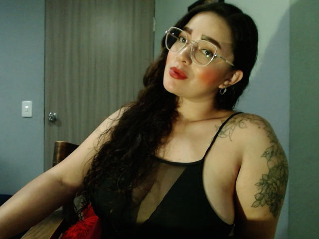 JaneAndPaull with curly dark hair, glasses, red lipstick, black sheer top, tattoos on right shoulder, looking over shoulder, seated, wooden door in background, slightly annoyed expression.