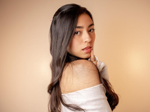 CanelleGarces with long dark hair, looking over shoulder, white off-shoulder top, finger touching mouth, brown eyes, soft smile, light skin, pink hairpin, gentle expression, warm lighting.