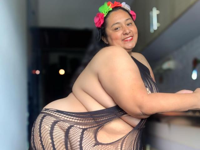 TistMature é uma mulher asiática corpulenta, com uma coroa de flores, sorrindo, vestindo um vestido preto de rede de peixe que revela seu grande e arredondado bumbum e seios. Ela está apoiada em uma bancada de cozinha..