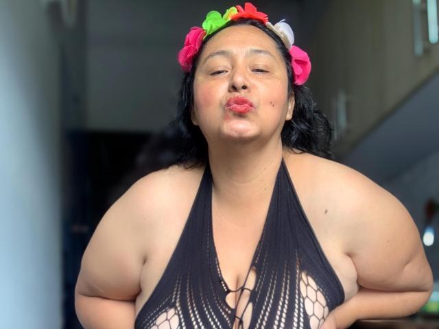 TistMature, une femme corpulente à la peau foncée et aux cheveux noirs et bouclés, porte un serre-tête fleuri et un haut en filet noir à encolure dégagée qui révèle ses seins volumineux, tout en pouting ses lèvres..