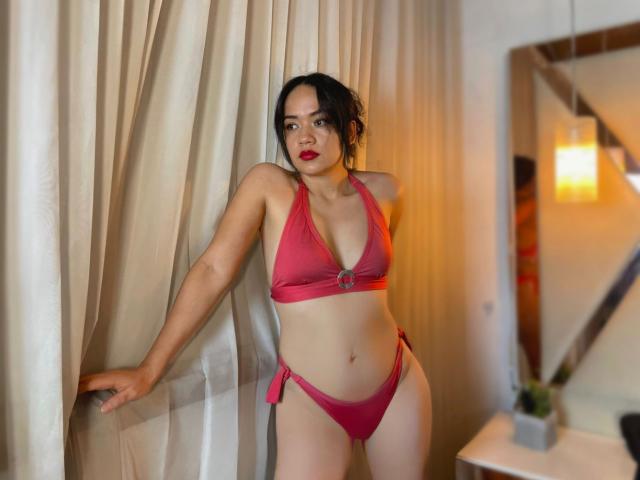 LorenXStar con un bikini rosa, de pie con una mano sobre cortinas blancas. Lleva lápiz labial rojo, cabello negro recogido en un moño despeinado, y una figura delgada y curvilínea con senos pequeños..