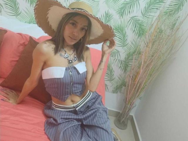 Veroo69 with tan skin, small tits, wearing a striped crop top, matching pants, and a big straw hat, sitting on a red bed, holding her hat.