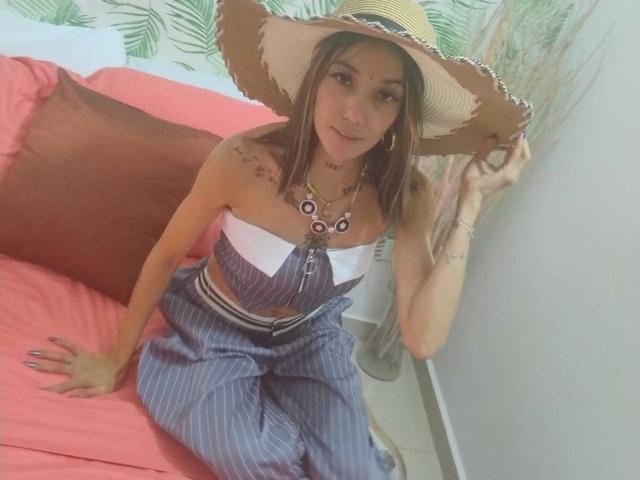 Veroo69 with tits in a white strapless top, wearing a big straw hat, pinstriped pants, and a necklace, sitting on a pink bed, holding her hat.