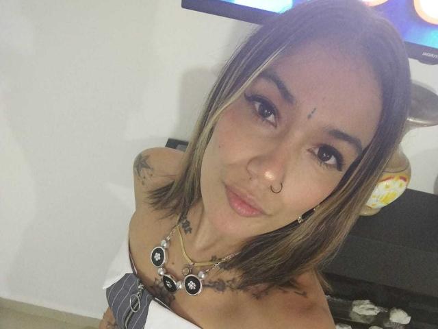 Veroo69 with brown and blonde hair, nose ring, tattoos, and necklace, looking up with pouty lips, holding white and gray striped cloth over small tits.