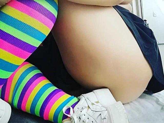 Mias bunte, gestreifte Socken und weiße Turnschuhe, kombiniert mit ihrer großen, nackten, blassen und schwarzen Shorts, während sie auf einem Bett sitzt. Die Socken haben leuchtend grüne, gelbe, blaue, pinke und violette Streifen..