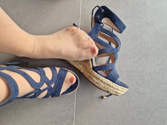 LilyStockings-hot's foot with pink toenails is touching a blue, woven-strap, high-heeled sandal on a gray tile floor. The other sandal is on her foot.