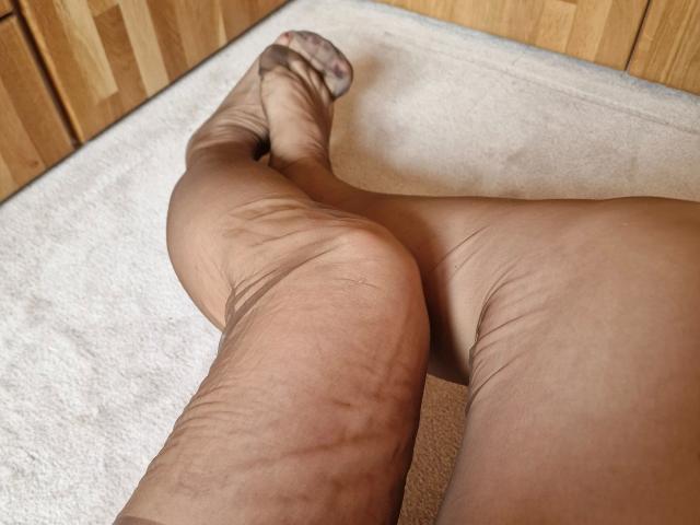 LilyStockings-hot's legs in sheer brown stockings, feet crossed, showing wrinkles and creases on her calves and toes, on a beige carpet in front of wooden cabinets.