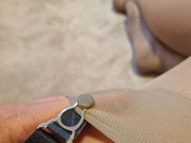 LilyStockings-hot's hand with a silver bracelet hooking into sheer beige stockings, showing the fabric's texture and slight stretch. Her toes are in the blurred background.