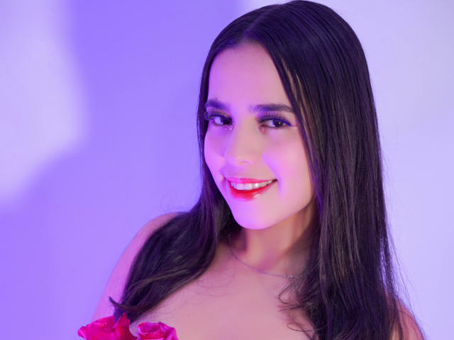 AlanaReyes with long black hair, red lipstick, smiling, purple light, pink rose on her chest, bare shoulders, silver necklace, looking at camera.