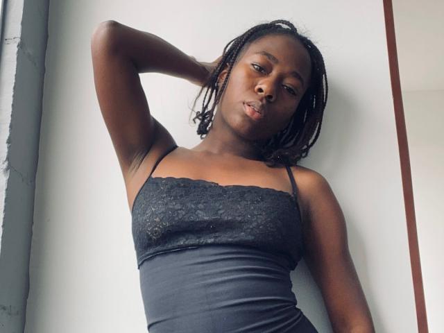 VioletGraice has dark skin, small tits, black lace spaghetti top, and black braids, standing with one arm behind her head, looking down with a slightly pouty expression.