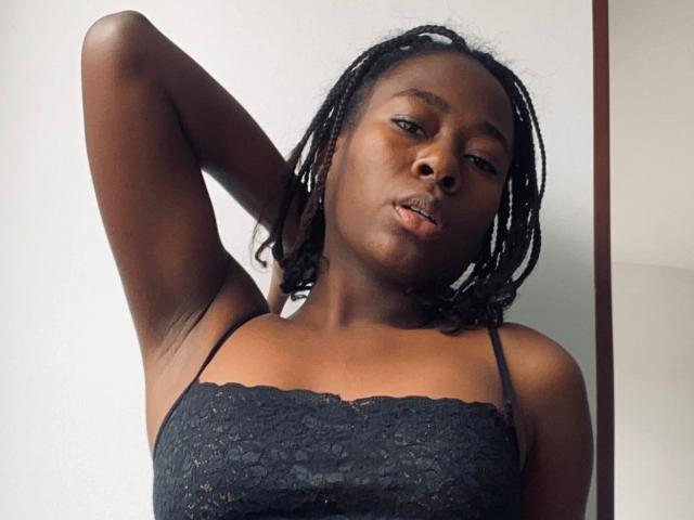 VioletGraice has dark skin, black lace top, and small breasts. She's looking at the camera with one arm raised, and her black dreadlocks are shoulder-length.