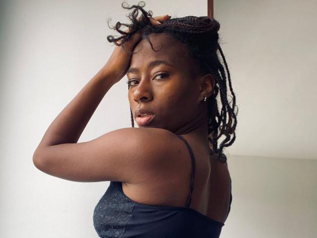 VioletGraice with dark brown skin, small round nose, and curly black dreadlocks, wearing a black spaghetti-strap top, hand in hair, looking over shoulder with a slight pout.