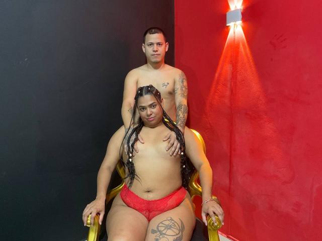 CamileXBastien, nue par le haut avec de grosses fesses, est assise sur une chaise dorée, vêtue de culottes en dentelle rouge. Elle a de longs cheveux tressés et des tatouages, tandis qu'un homme tatoué et sans chemise se tient derrière elle..