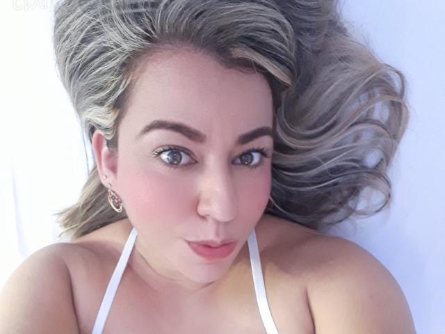 AnmeLeon has light skin, gray-green eyes, and pink blush on her cheeks. She's wearing a white halter top, with curly gray-blonde hair and small, ornate earrings.