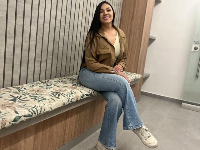 YessiJaymes seduta su una panchina di legno con un cuscino a motivi floreali, indossa una giacca marrone, jeans blu e scarpe da ginnastica bianche, sorride con lunghi capelli neri, seno di media grandezza..