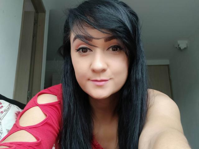Katnnis with black hair, brown eyes, and pink lipstick, wearing a red top with cutout sections, showing her tits slightly, taking a selfie, smiling softly.