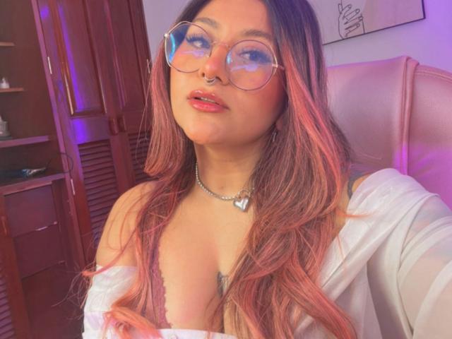 LorraineV with pink-tinted hair, glasses, nose ring, and white top showing cleavage, wearing a heart necklace, sitting in a pink-lit room. Boobs visible, slightly parted lips.