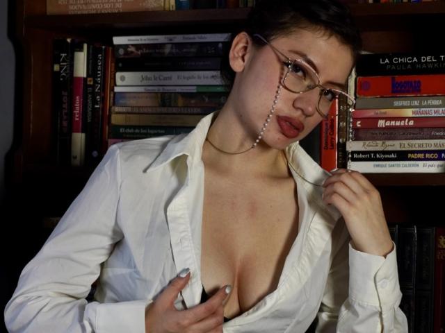 ElizaCanterville with short brown hair, glasses, and red lipstick, wearing an unbuttoned white shirt, exposing her small tits, holding her shirt open, standing in front of bookshelves.