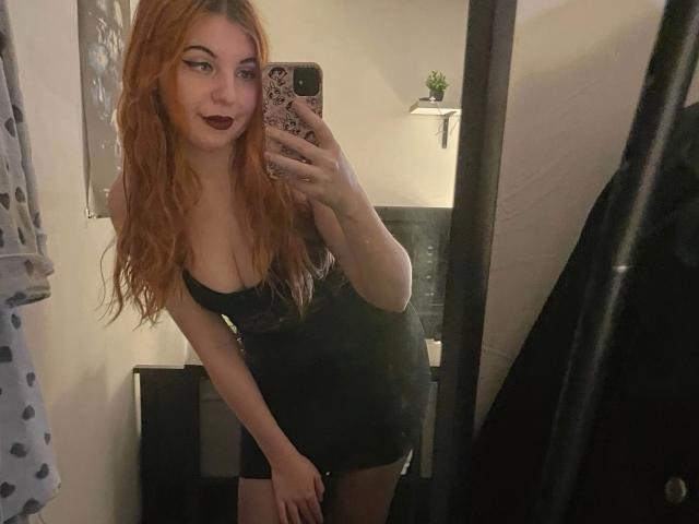 Luna-Noir-hot with red hair, dark lipstick, and black dress, showing her tits in a mirror selfie, holding a phone with a pink case. Her ass is slightly visible.