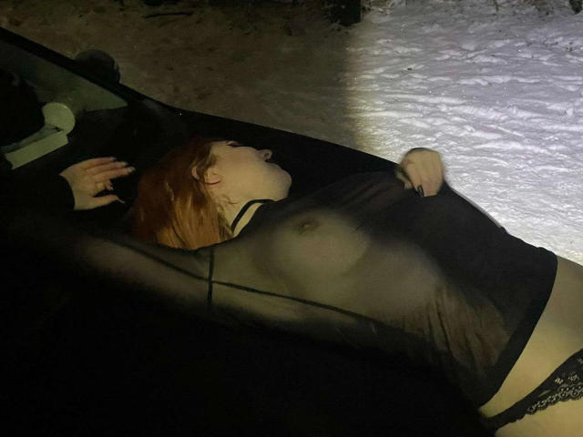 Luna-Noir-hot, with red hair, is lying in a black car, wearing a sheer black top, exposing her tits and black lace panties, in a snowy night scene.