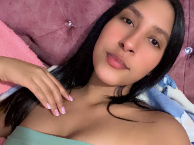 MadisonReyes has light brown skin, black hair, and pink nails. She's wearing a green strap top, lying on pink and blue blankets, with a pink, tufted headboard.