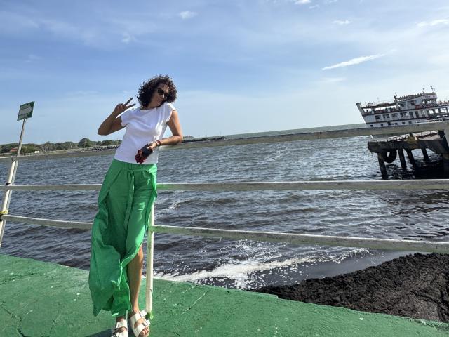 MatureJennyX with curly dark hair, wearing a white top and green flowing skirt, standing on a green dock, holding a red bottle, and looking at the camera.