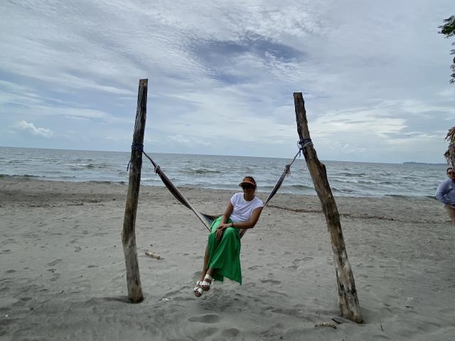 MatureJennyX, white shirt, green skirt, brown cap, hanging from wooden swing, over sandy beach, ocean in background, cloudy sky, wearing white sandals, relaxed, casual, slight smile.