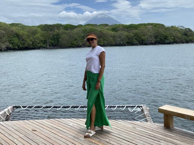 MatureJennyX stands on a wooden dock, wearing a white tee, green skirt with a thigh slit, brown hat, sunglasses, and white sandals. Mountain and trees in the background.