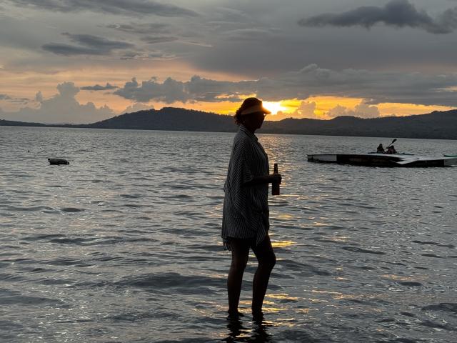 MatureJennyX stands in water, wearing a striped shawl, holding a drink, sunset in background, small boat to right, clouds and orange sky, silhouette, water reflects light.