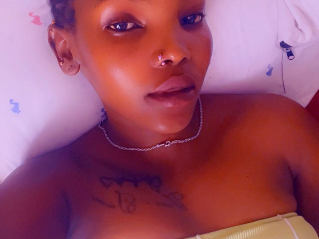SexxyMayana with dark skin, nose ring, and chain necklace, lying down, showing bare shoulders and chest tattoo, looking at camera with slightly parted lips, purple-tinted photo.