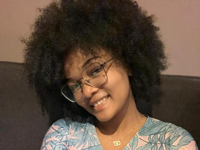 Lulucahs with a big, curly black afro, wearing glasses and a blue-patterned top, smiling slightly, has a small gold necklace with a crescent moon pendant.