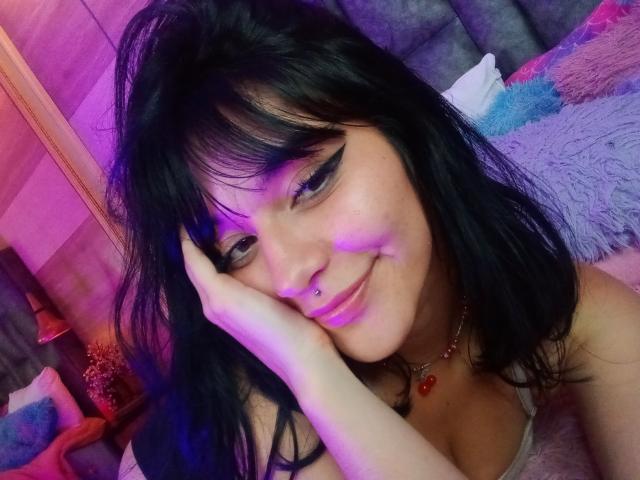 LindaWatsong with black hair, eyeliner, nose piercing, smiling, hand on cheek, purple and pink lighting, necklace with red gem, visible cleavage in pink top, looking at camera.