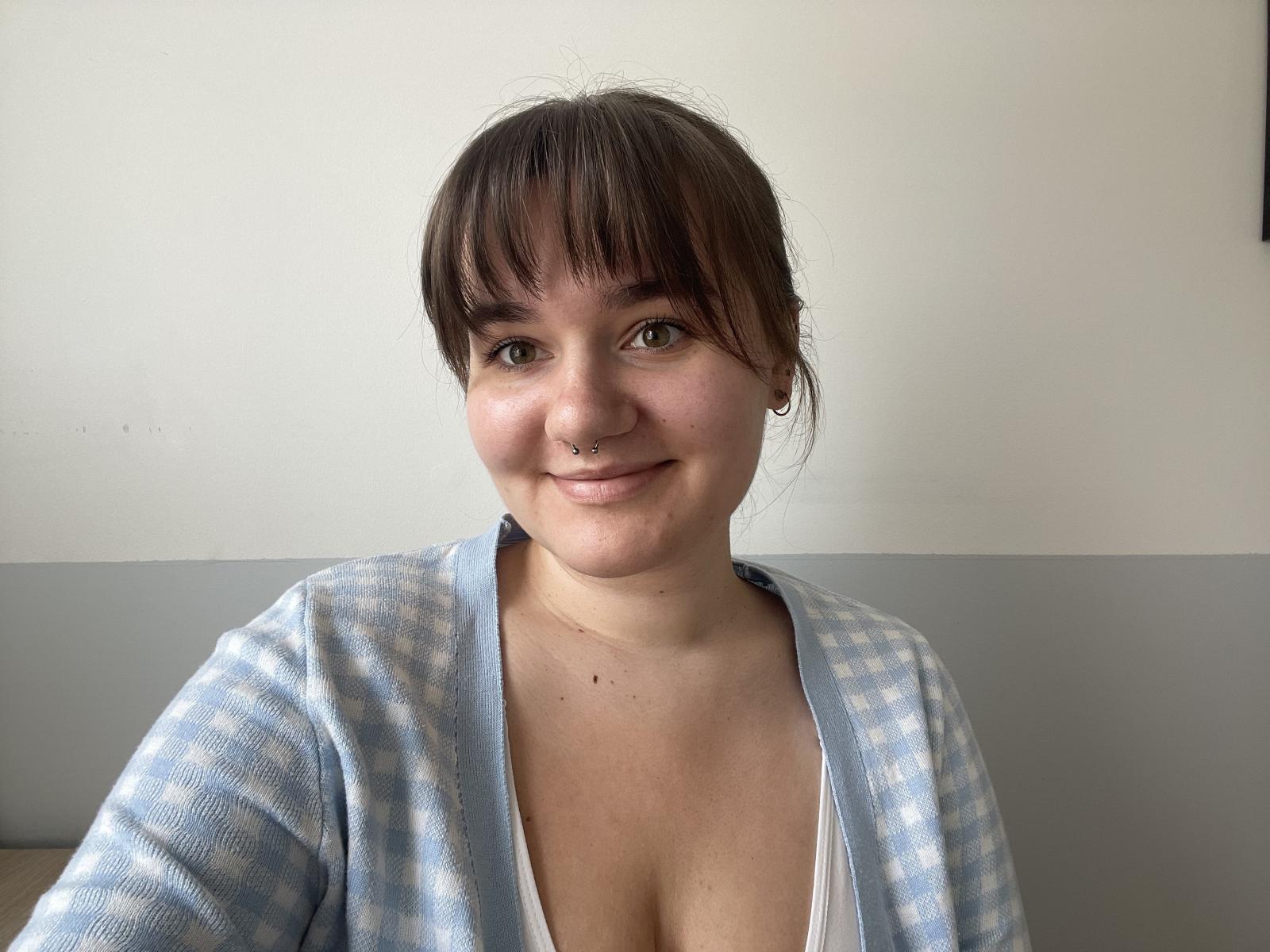 JulieTease with brown hair, bangs, and nose ring, smiling, wearing a blue checkered cardigan, exposing her cleavage, and a white shirt, in a selfie.