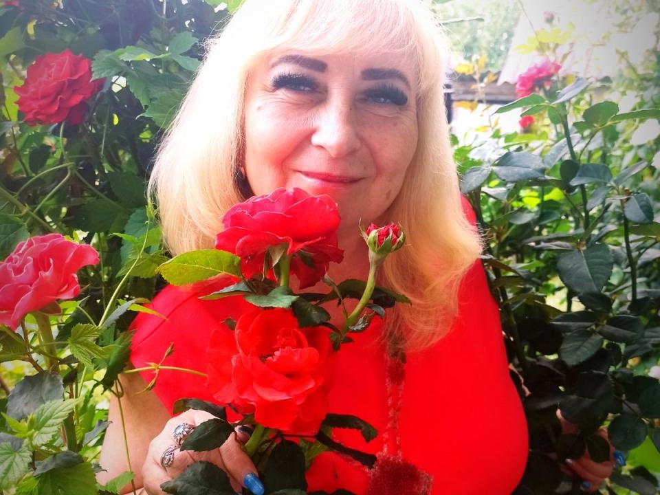 AlinaXX69 with blonde hair, wearing a red top, holding red roses, smiling, her tits slightly visible, surrounded by green leaves and more red roses.